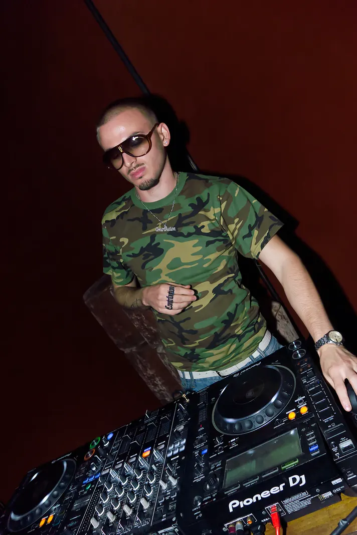 Berjyy behind a Pioneer DJ deck, hand on his chest, tilted low-angle shot