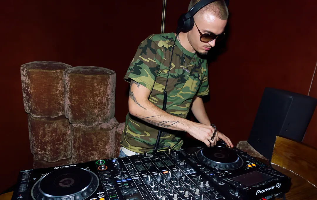 Berjyy mixing on a Pioneer DJ setup, headphones on, against a dark red wall